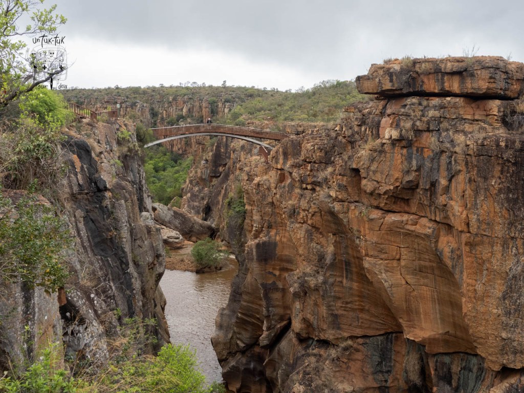Vue sur un canyon profond avec un ruisseau et un pont en arc en arrière-plan, entouré de formations rocheuses et de végétation.
