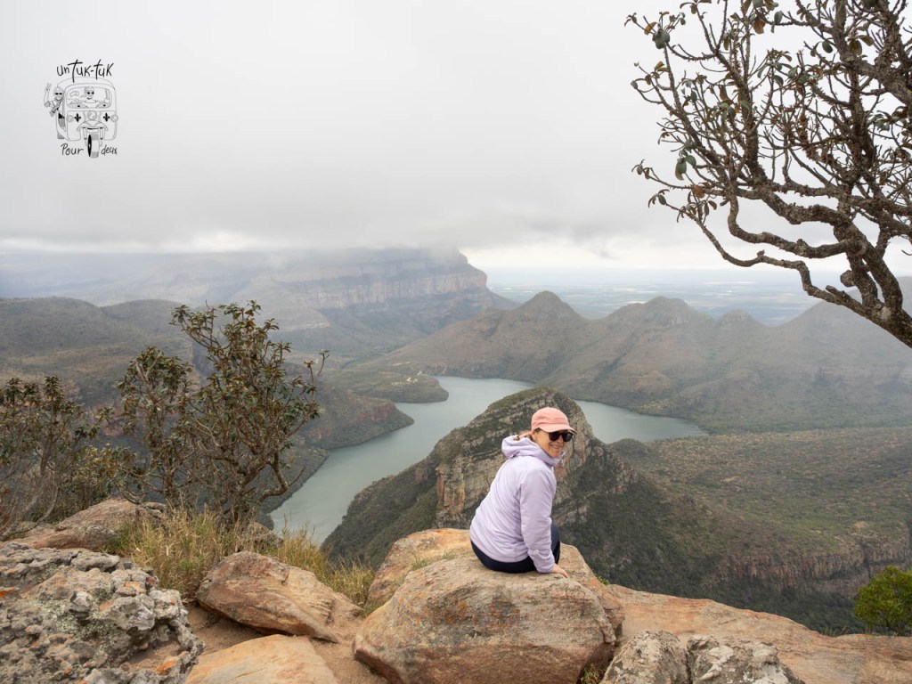 Une personne assise sur une roche au bord d'une falaise, avec une vue panoramique sur un paysage montagneux et un lac en contrebas, sous un ciel nuageux.