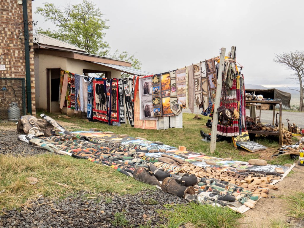 Un marché en plein air avec des tissus colorés accrochés, des objets artisanaux exposés au sol, et un bâtiment à l'arrière-plan.