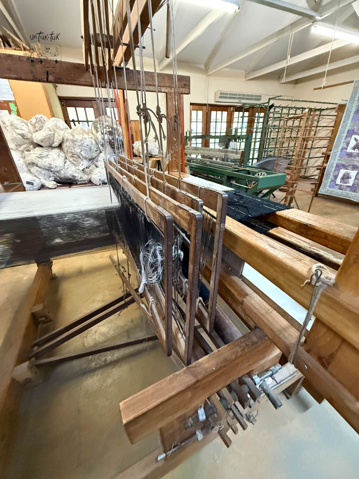 Métier à tisser en bois dans un atelier, avec des fils de trame visibles et des rouleaux de textile en arrière-plan.