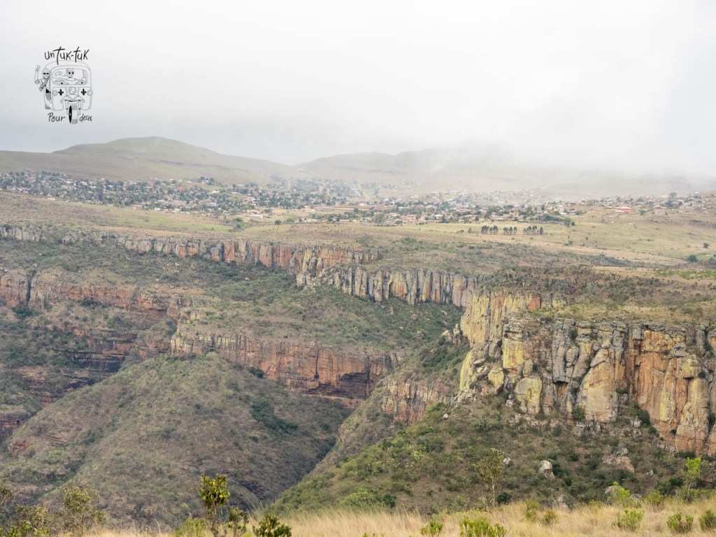 Vue panoramique d'un canyon avec des formations rocheuses, au loin une ville sur une colline sous un ciel nuageux.