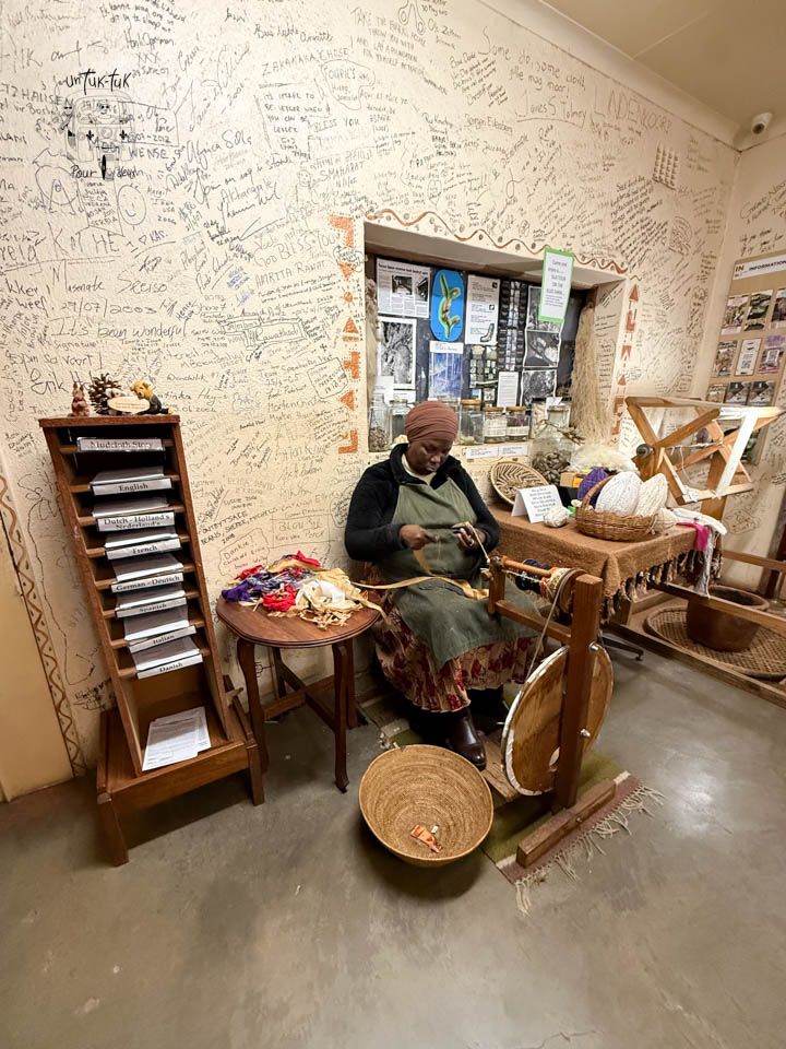 Une femme assise à une roue de filage, en train de travailler des fibres colorées, dans une pièce avec des murs couverts d'inscriptions et de notes.