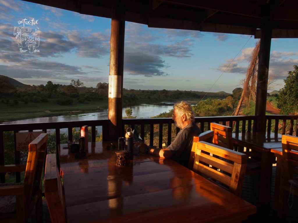 Un homme assis à une table en bois surplombant un paysage paisible avec un fleuve et des collines au coucher du soleil.