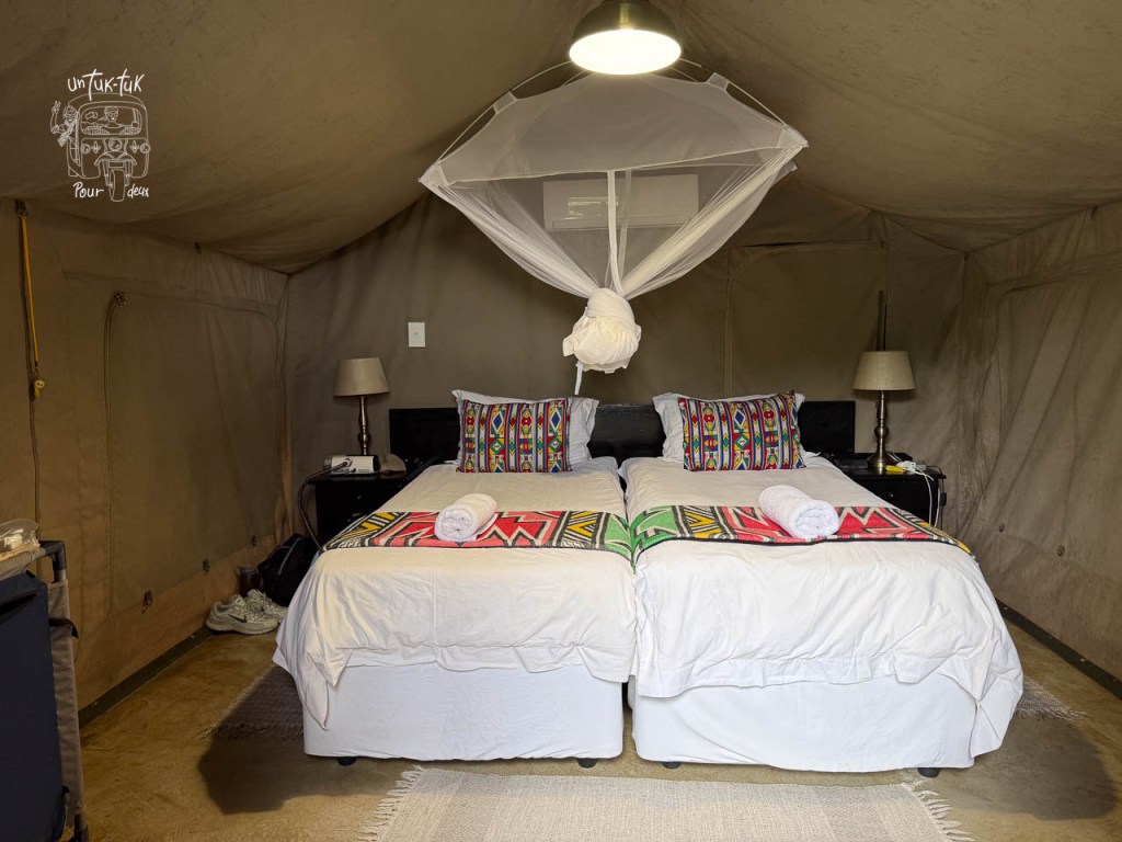 Chambre intérieure d'un hébergement avec deux lits jumeaux, des draps blancs, des coussins colorés et une moustiquaire au-dessus des lits. Éclairage doux avec lampes de chevet et décoration dans un magasin en toile.
