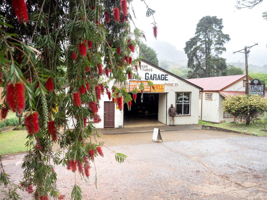 Un garage avec un homme à l'entrée, entouré de fleurs rouges et d'arbres dans une atmosphère pluvieuse.