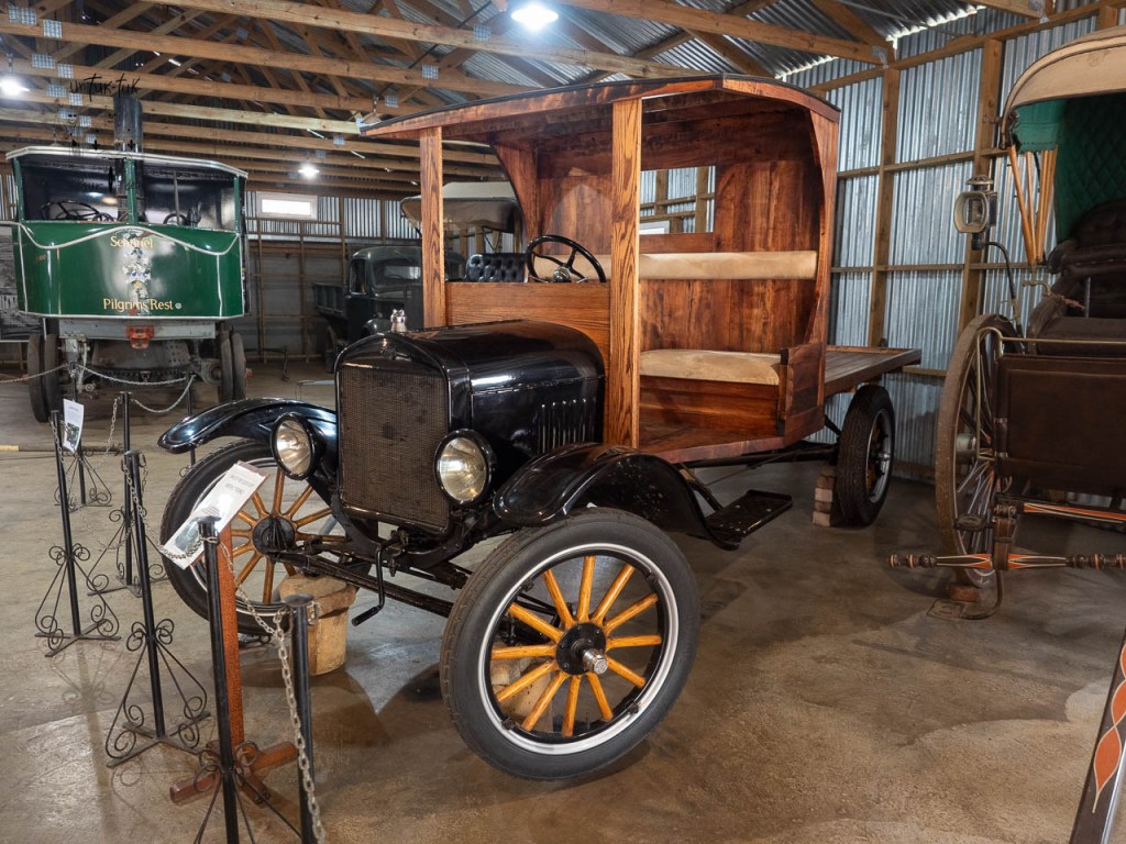 Un ancien camion noir en bois exposé dans un musée, avec des roues à rayons et un compartiment ouvert à l'arrière.