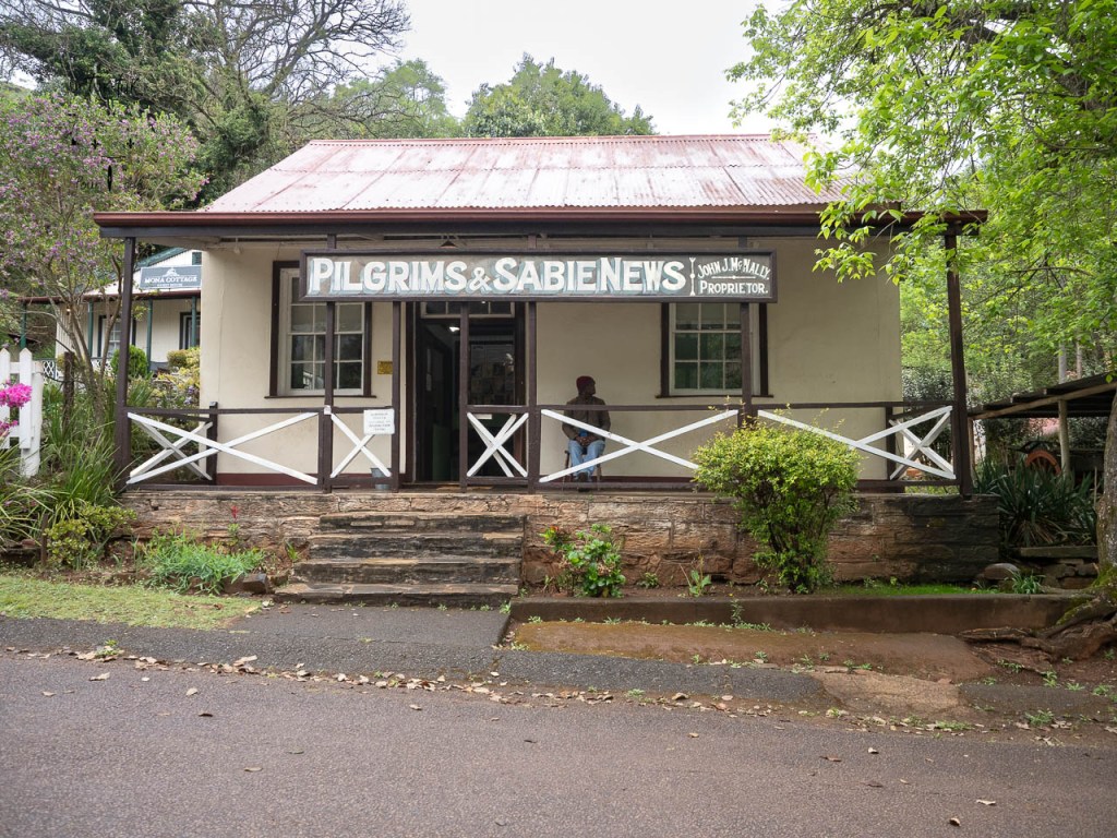 Façade d'un bâtiment nommé 'Pilgrims & Sabie News' avec un propriétaire visible sur le porche, entouré de verdure.